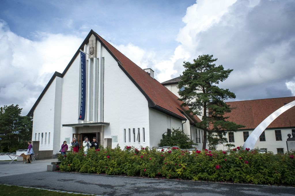 녹색으로 둘러싸인 빨간색 지붕과 "Vigelandmuseet" 배너가 있는 흰색 교회 같은 건물과 입구에는 여러 사람이 있습니다.
