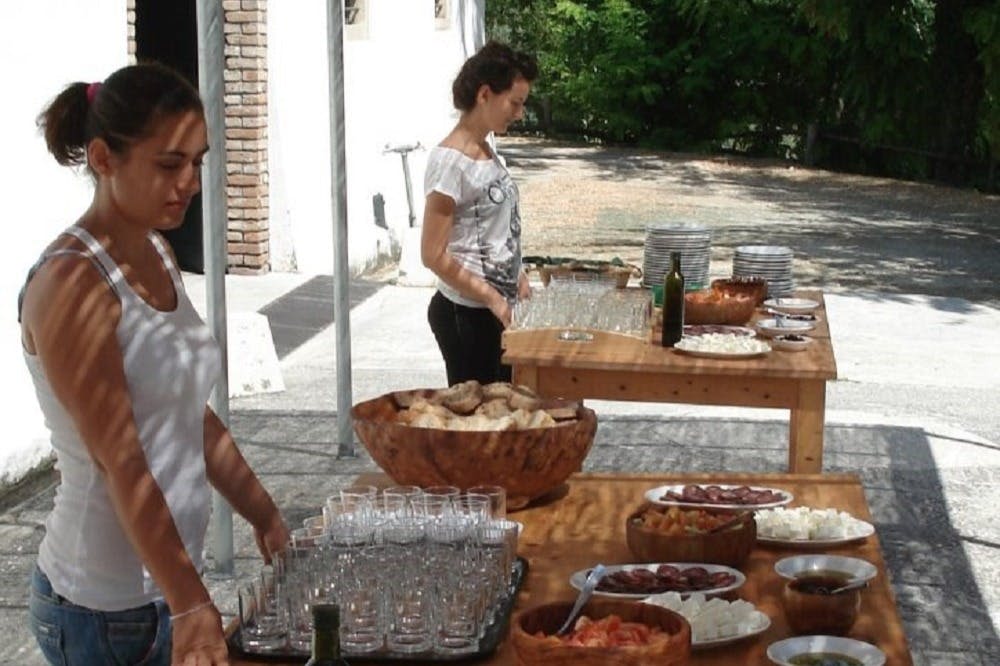 흰색 건물과 녹지 근처의 야외 공간에서 두 명의 여성이 빵과 여러 잔이 놓인 테이블을 준비합니다.