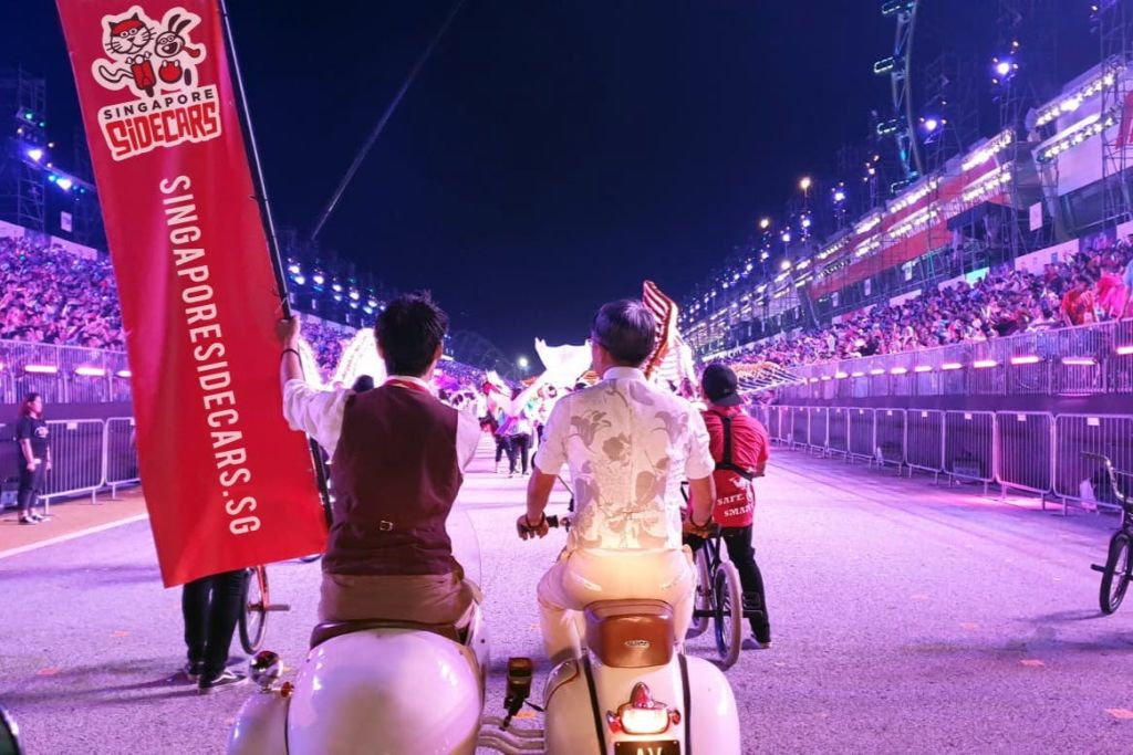 밤에 조명이 켜진 거리에서 빈티지 사이드카와 자전거를 타고 빨간색 배너를 들고 있는 두 사람. 군중은 백그라운드에서 볼 수 있습니다.