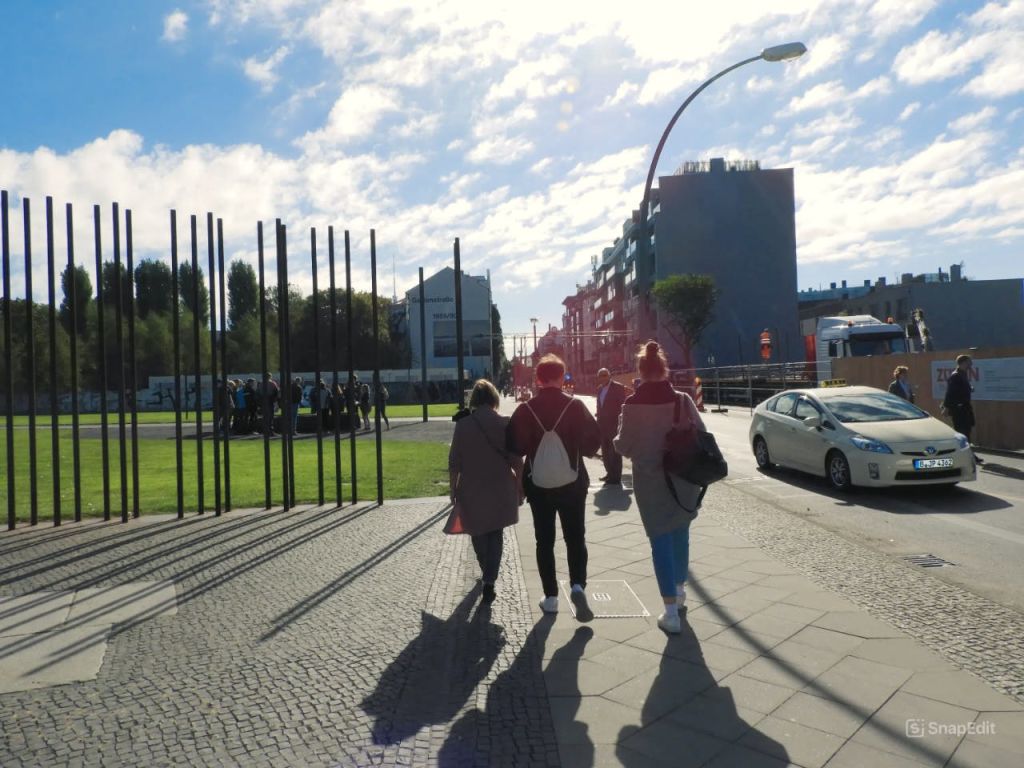 건물과 굴곡진 가로등을 배경으로 잔디밭 근처의 햇살 가득한 보도를 걷고 있는 세 사람.