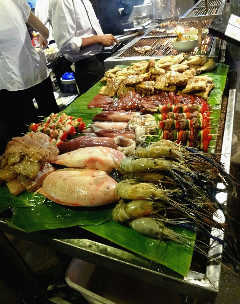 다양한 해산물, 고기, 야채 꼬치가 푸드 마켓 가판대의 푸른 잎에 전시되어 있습니다. 두 사람이 뒤에서 구워먹고 있습니다.