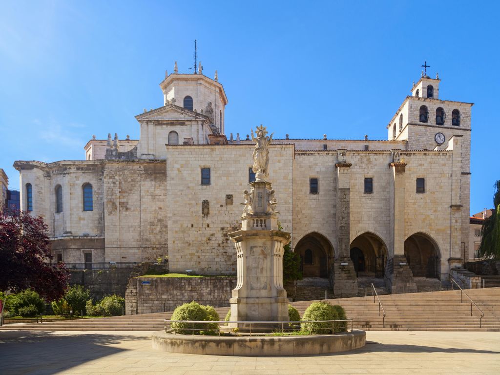 아치형 입구가 있는 대형 석조 성당, 조각상이 있는 중앙 분수대, 잘 다듬어진 수풀이 있는 맑고 푸른 하늘 아래 있습니다.