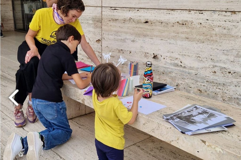 종이, 마커, 장식품을 비롯한 다채로운 소모품이 놓인 테이블에서 두 명의 어린이와 한 명의 어른이 공예품을 만들고 있습니다.