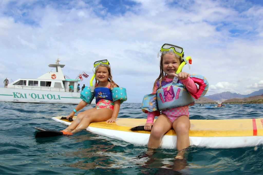 스노클링과 구명조끼를 입은 두 어린이가 바다에서 패들보드에 앉아 부분적으로 흐린 하늘 아래 미소를 짓고 있습니다.