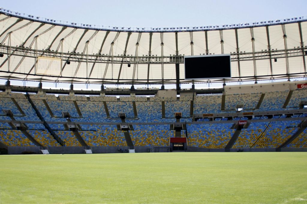 파란색과 노란색 좌석, 대형 점수판, 부분적으로 덮인 지붕 아래 녹색 필드가 있는 빈 경기장.