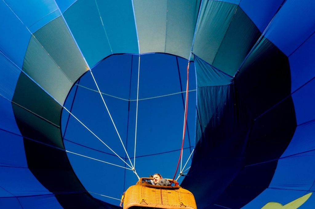 열기구 바구니에 서서 맑고 푸른 하늘을 배경으로 커다란 파란색과 청록색 풍선을 올려다보고 있는 사람.