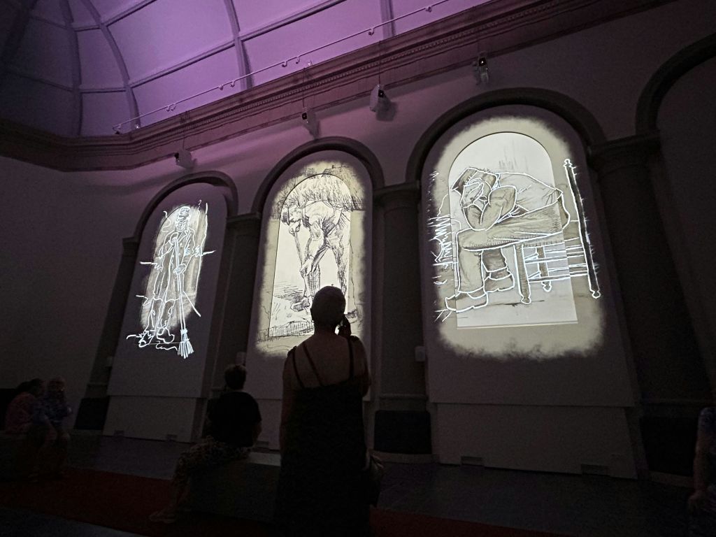 아트 갤러리의 세 개의 높은 아치형 패널에 투영된 크고 조명된 스케치를 보는 사람들.