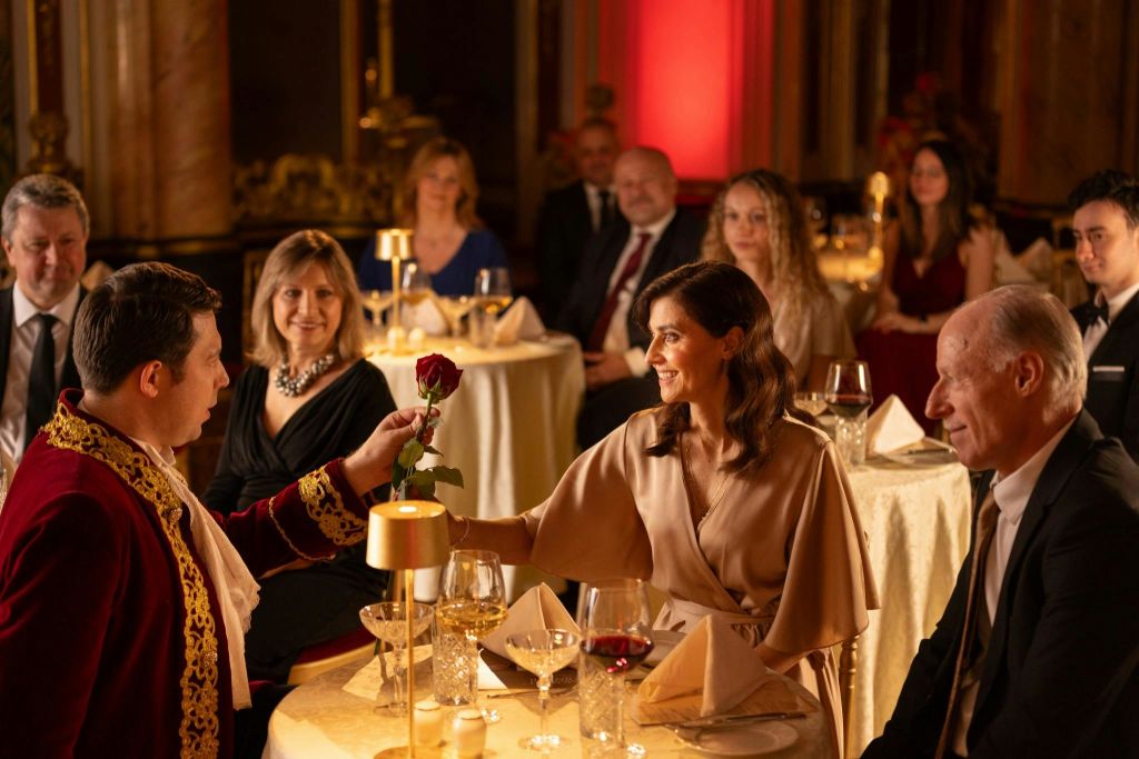 레드 벨벳 재킷을 입은 한 남성이 우아하게 차려입은 손님들과의 정찬에서 미소 짓는 여성에게 빨간 장미를 선물합니다.