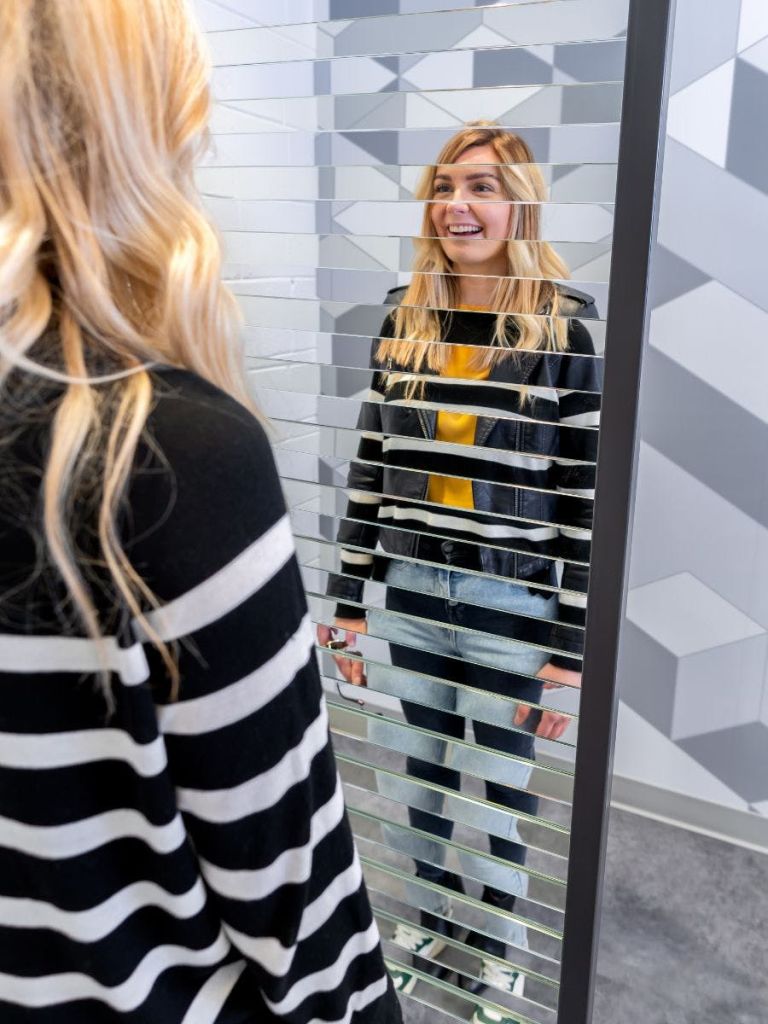 한 명은 줄무늬 셔츠를 입고 한 명은 검은색 재킷과 노란색 셔츠를 입은 두 여성이 줄무늬 유리 칸막이를 통해 서로 마주보고 있습니다.