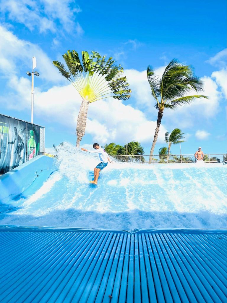 한 사람이 야자수를 배경으로 밝고 맑은 하늘 아래 푸른 인공 파도를 타고 서핑을 하고 있습니다.