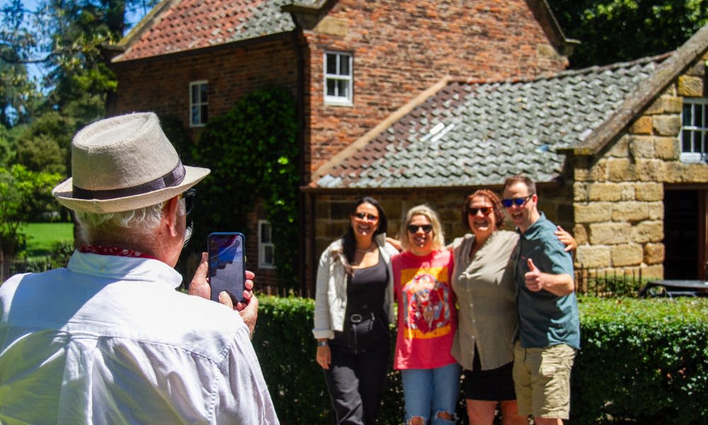 화창한 날 타일로 마감된 지붕이 있는 석조 건물 앞에서 웃으며 팔짱을 끼고 있는 네 사람.