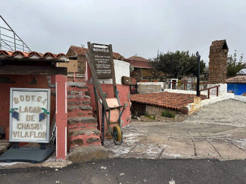 벽돌 벽, 오래된 손수레, 석조 계단, 보데가 표지판이 있는 소박한 풍경. 타일로 덮인 지붕과 흐린 하늘을 배경으로.