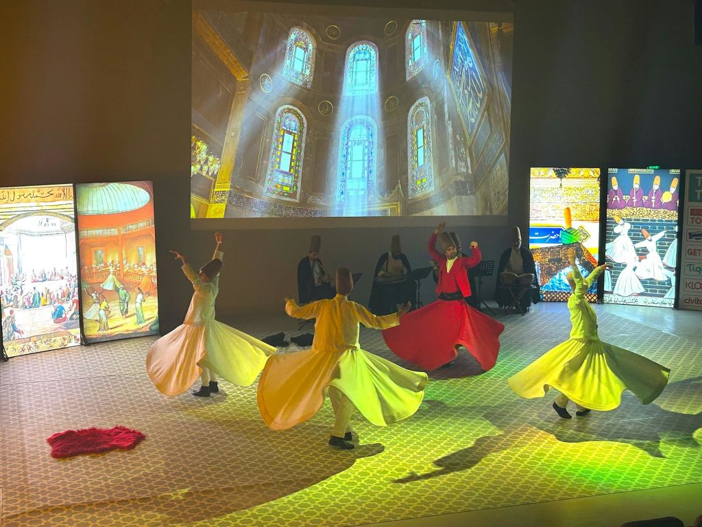 화려한 회오리 의상을 입은 댄서들이 조명이 희미한 방에서 영사된 이미지를 배경으로 공연을 펼칩니다.