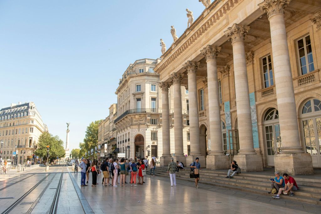 한 무리의 사람들이 지붕 위에 기둥과 조각상이 있는 웅장한 건물 밖에 서서 맑고 푸른 하늘을 감상하고 있습니다.