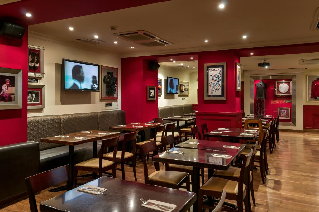 빨간색 벽, 액자 예술, TV 화면, 나무 테이블, 의자가 깔끔하게 배치된 레스토랑 인테리어입니다.