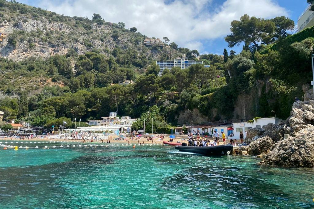 해변, 푸른 언덕, 건물, 부분적으로 흐린 하늘 아래 청록색 물 위에 정박한 보트가 있는 해안 풍경.