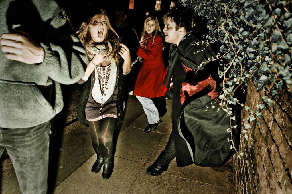 뱀파이어 옷을 입은 남자가 어두운 거리에서 두 여자를 겁주는 척합니다. 한 여자는 과장된 두려움을 보입니다.