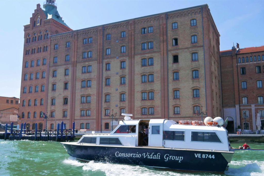 "콘소르치오 비달리 그룹 (Consorzio Vidali Group)" 이라고 쓰여진 보트가 대형 복층 건물 앞에서 물 위를 여행합니다.