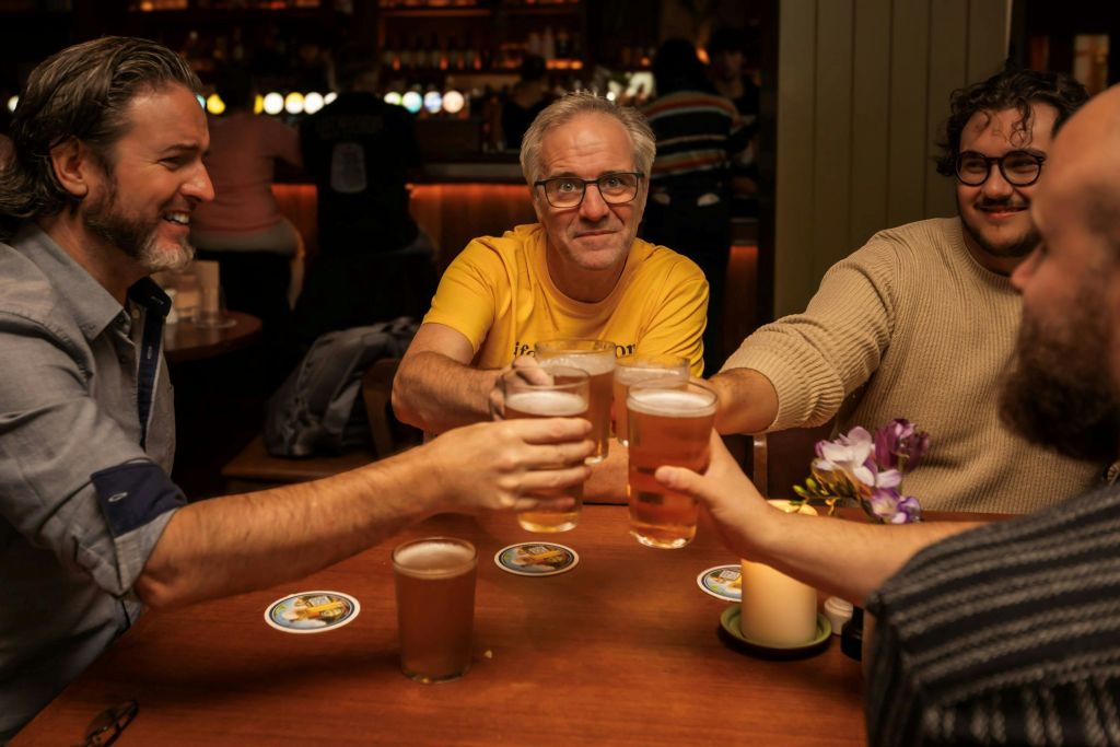 한 무리의 사람들이 아늑한 분위기의 바에서 맥주 한 잔을 마시며 건배하고, 노란 셔츠를 입은 한 남자가 미소를 짓고 있습니다.