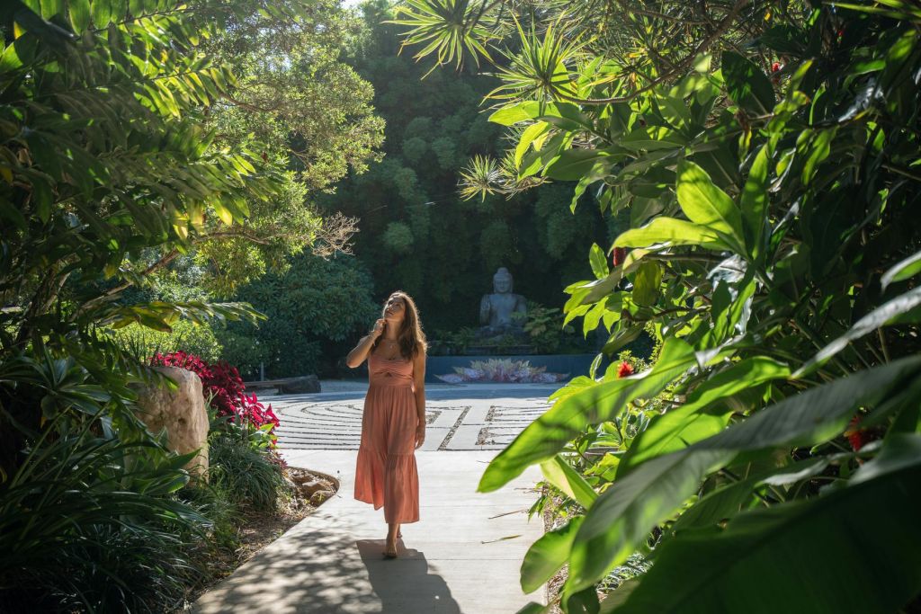 산호색 드레스를 입은 한 여성이 녹음이 우거진 정원길을 따라 걸어가고 있으며, 배경에는 불상이 있습니다.