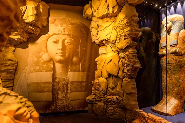 박물관을 배경으로 암석 지층을 닮은 질감의 벽으로 둘러싸인 고대 인물의 얼굴이 담긴 대형 문화 예술 작품입니다.