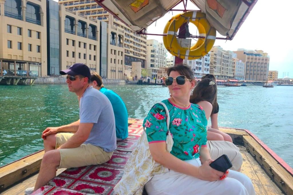 세 사람이 보트에 앉아 무늬가 있는 바닥 매트와 건물, 선착장을 배경으로 물놀이를 즐기고 있습니다.