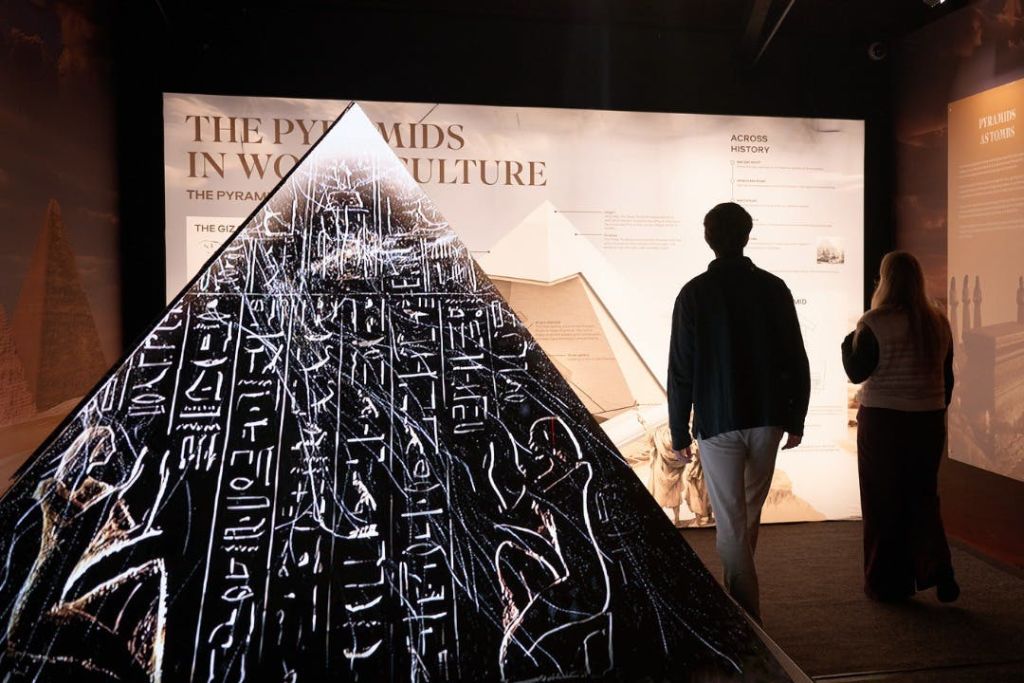 한 사람이 피라미드의 역사에 대한 전시 벽 앞에서 상형 문자가 있는 불이 켜진 피라미드 모양의 디스플레이를 관찰합니다.