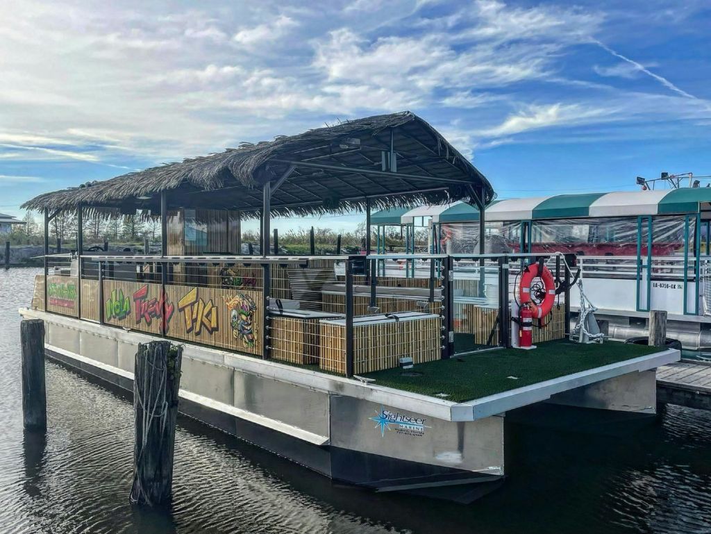 초가 지붕, 야외 좌석, 다채로운 색상의 간판이 있는 수상 티키 바가 잔잔한 물가에 정박해 있습니다.