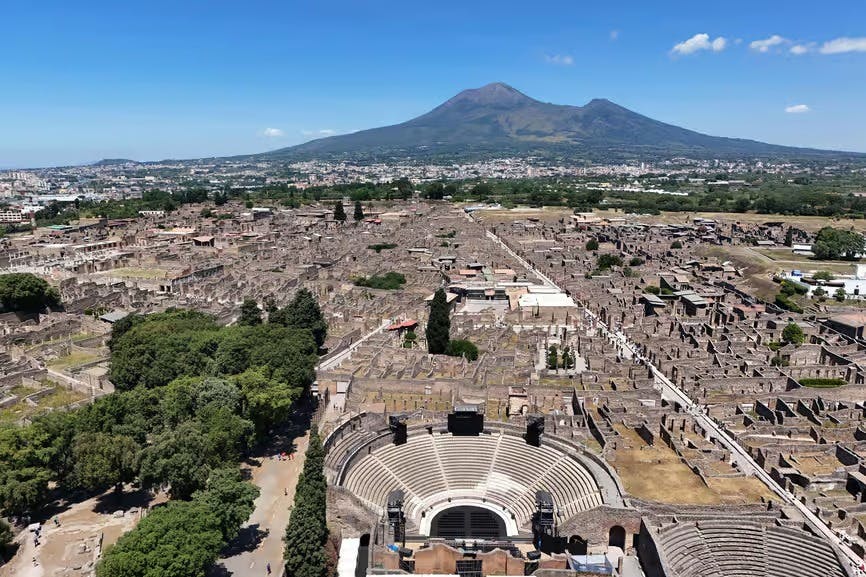 전경의 원형 극장과 푸른 나무, 그리고 베수비오 산을 배경으로 폼페이 유적의 공중 전경을 감상할 수 있습니다.
