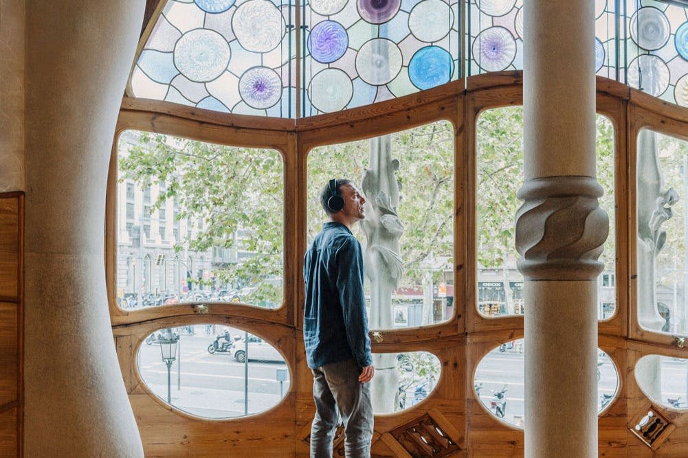 나무, 자동차, 건물이 있는 번화가가 내려다보이는 크고 장식적인 창문 근처에 헤드폰을 착용한 사람이 서 있습니다.