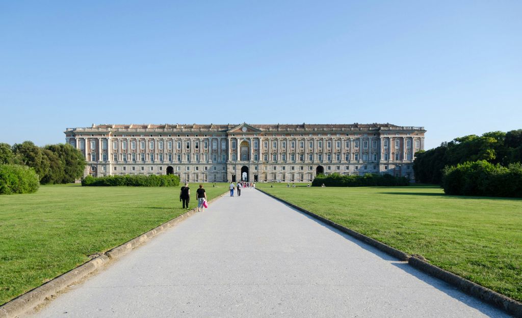 커다란 유서 깊은 궁전으로 이어지는 넓은 길을 걷는 사람들은 여러 개의 창문, 화려한 디테일, 잘 손질된 잔디밭으로 둘러싸인 아치로 이어집니다.