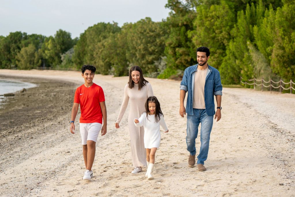4인 가족이 모래사장을 따라 산책합니다. 나무를 배경으로 야외 활동을 즐기며 미소를 짓고 있습니다.