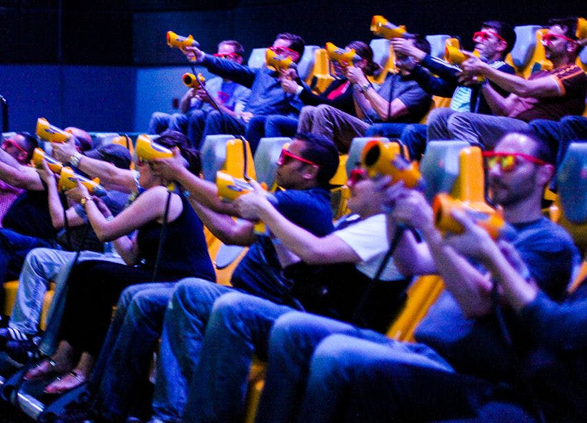 3D 안경을 쓰고, 노란 총을 들고, 인터랙티브한 어트랙션에서 스크린을 조준하는 사람들이 줄지어 앉아 있습니다.