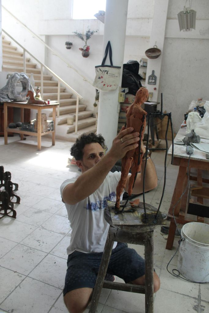 한 사람이 테이블, 도구, 계단을 배경으로 작업장에서 점토로 작은 사람의 모습을 조각하고 있습니다.