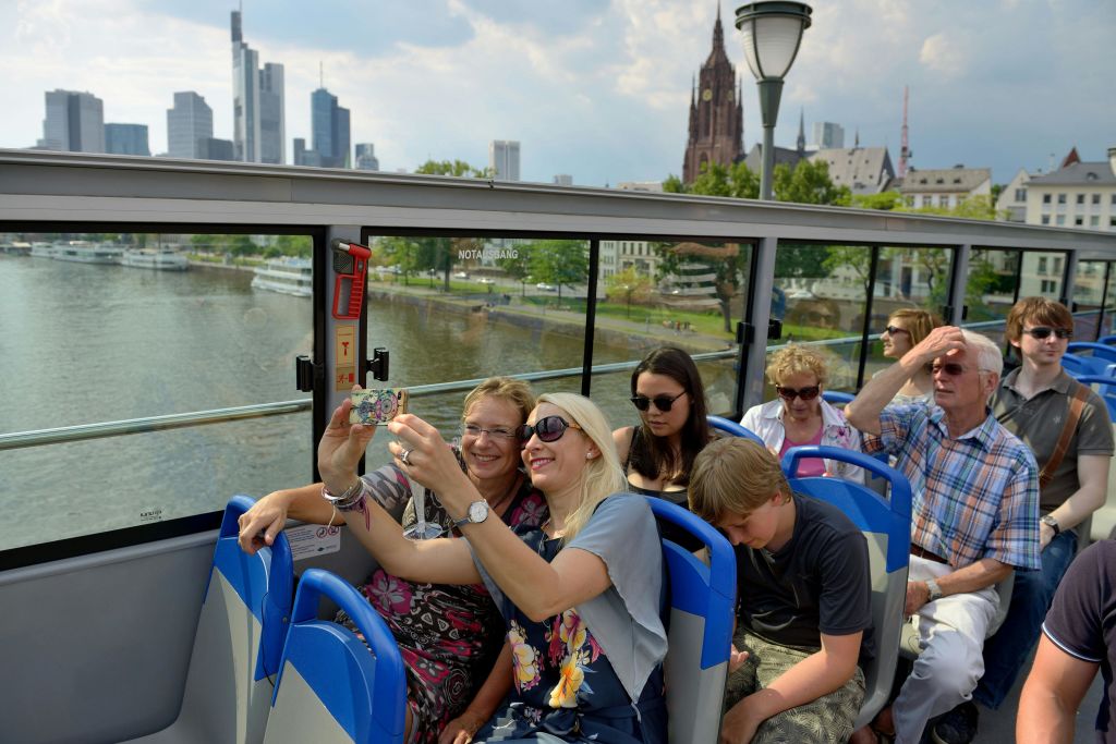 어린 소년과 셀카를 찍는 여성을 포함한 일행이 도시 풍경을 배경으로 오픈탑 투어 버스를 타고 있습니다.