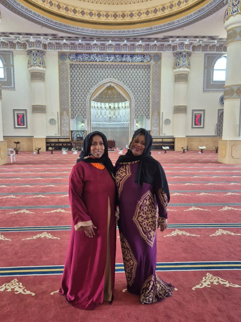 겸손한 드레스와 히잡을 입은 두 명의 여성이 정교한 아치와 레드 카펫 바닥이 있는 화려한 모스크 안에 서 있습니다.