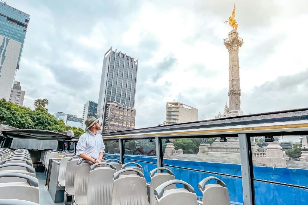 하얀 셔츠와 모자를 쓴 남성이 투어 버스에 서서 흐린 하늘 아래 도시 건물 사이에 있는 높은 기념비를 바라보고 있습니다.