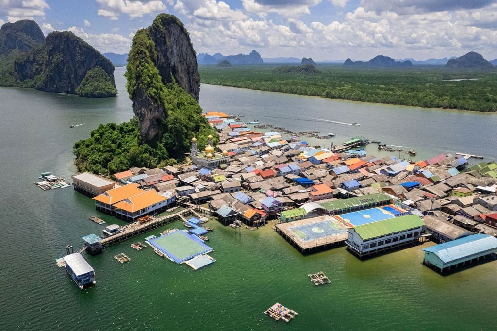 부분적으로 흐린 하늘 아래 물과 울창한 녹색 산으로 둘러싸인 다채로운 지붕이 있는 해안 마을의 공중 전망.