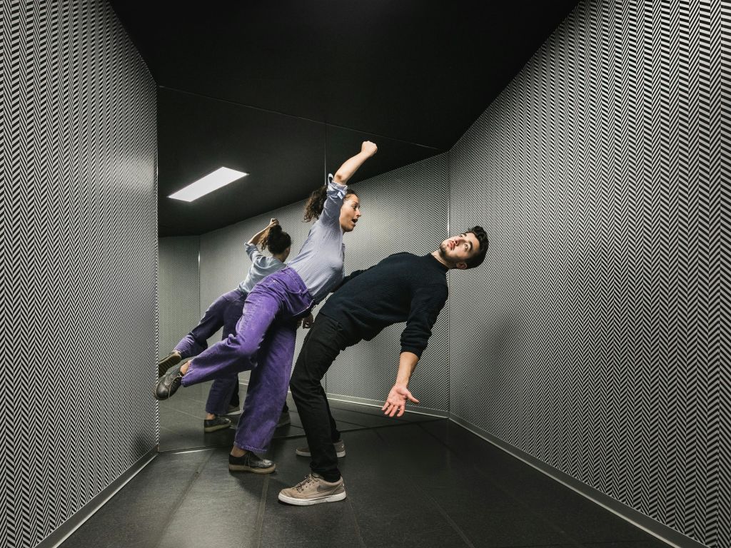 두 사람이 거울처럼 무늬가 있는 방에서 불가능한 각도로 몸을 기울이며 중력을 거스르는 것처럼 보입니다.