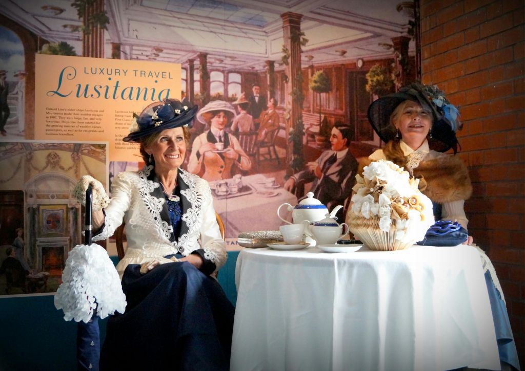 빈티지 복장을 한 두 명의 여성이 찻주전자와 장식품이 놓인 테이블에 앉아 있으며, 유서 깊은 식사 현장의 벽화를 감상할 수 있습니다.