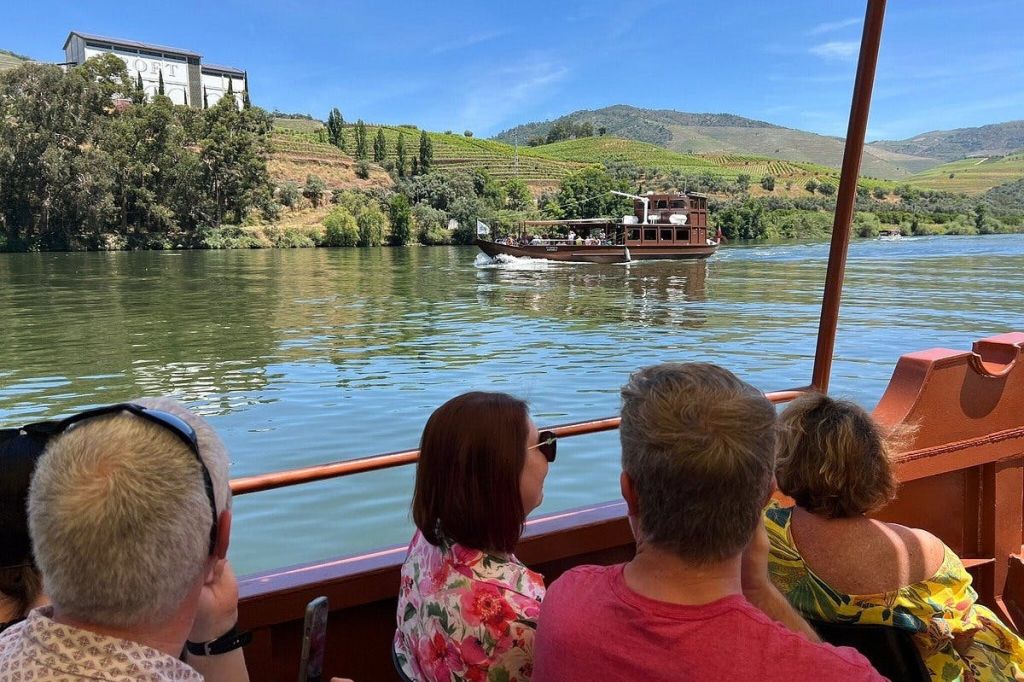 보트를 탄 사람들은 맑고 푸른 하늘 아래 포도원과 언덕을 배경으로 강에서 또 다른 보트를 바라보고 있습니다.