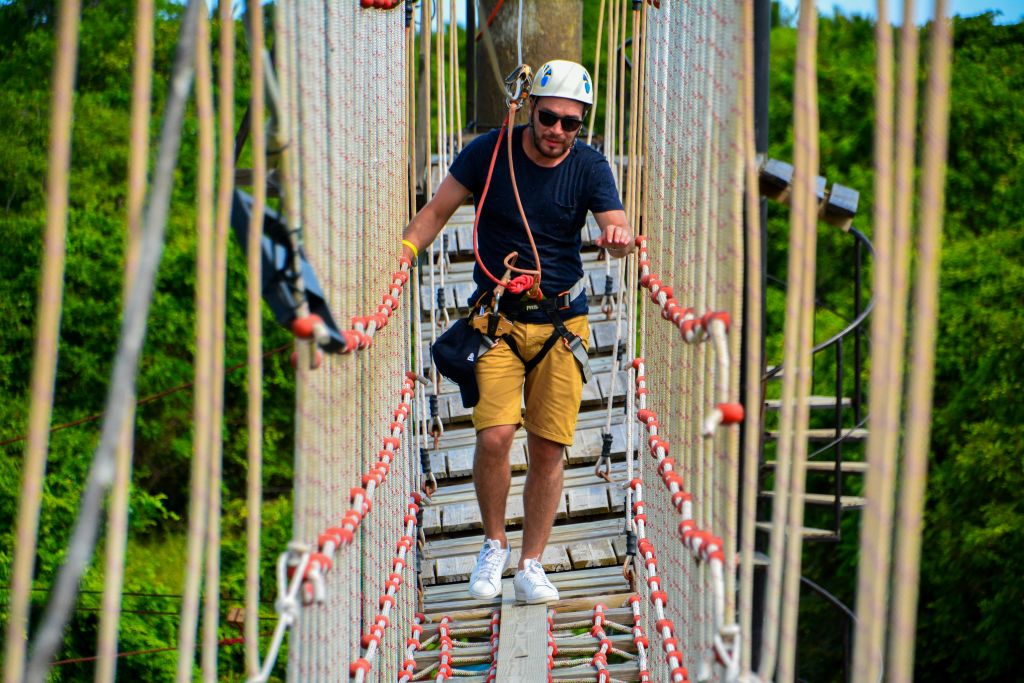 헬멧과 하네스를 쓴 남성이 울창한 푸른 나무에 둘러싸인 그물망이 달린 밧줄 다리를 걷고 있습니다.