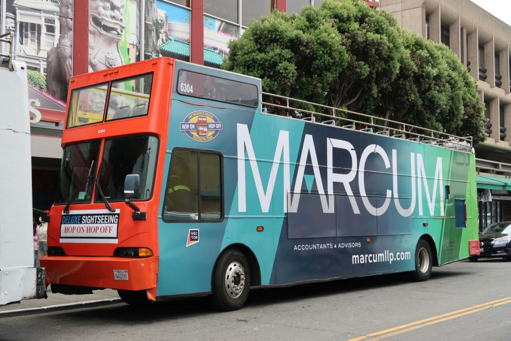 도시 거리에 주차된 "Marcum" 광고가 있는 2층 관광 버스. 나무와 건물이 배경입니다.