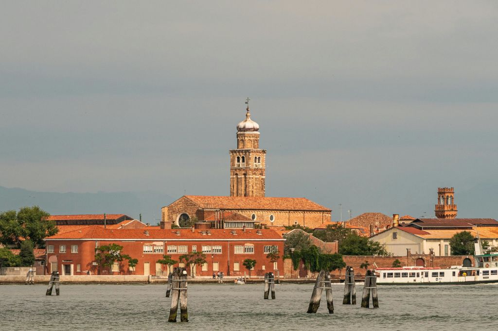 구름 낀 하늘과 잔잔한 물을 배경으로 돔형 지붕이 있는 높은 탑 위에 붉은 벽돌로 마감된 건물이 있는 해안 풍경.