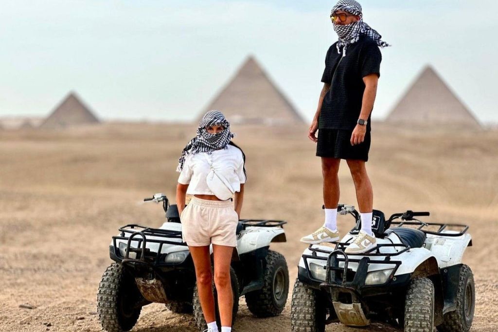 머리 스카프를 두른 두 사람이 모래 지형의 피라미드 앞 ATV에 서서 앉아 있습니다.