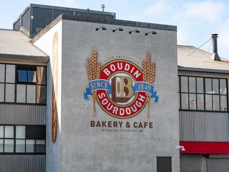 건물 벽에 칠해진 Boudin Sourdough Bakery & Cafe 로고는 밀 줄기, 텍스트, 커다란 "B" 엠블럼을 특징으로 합니다.