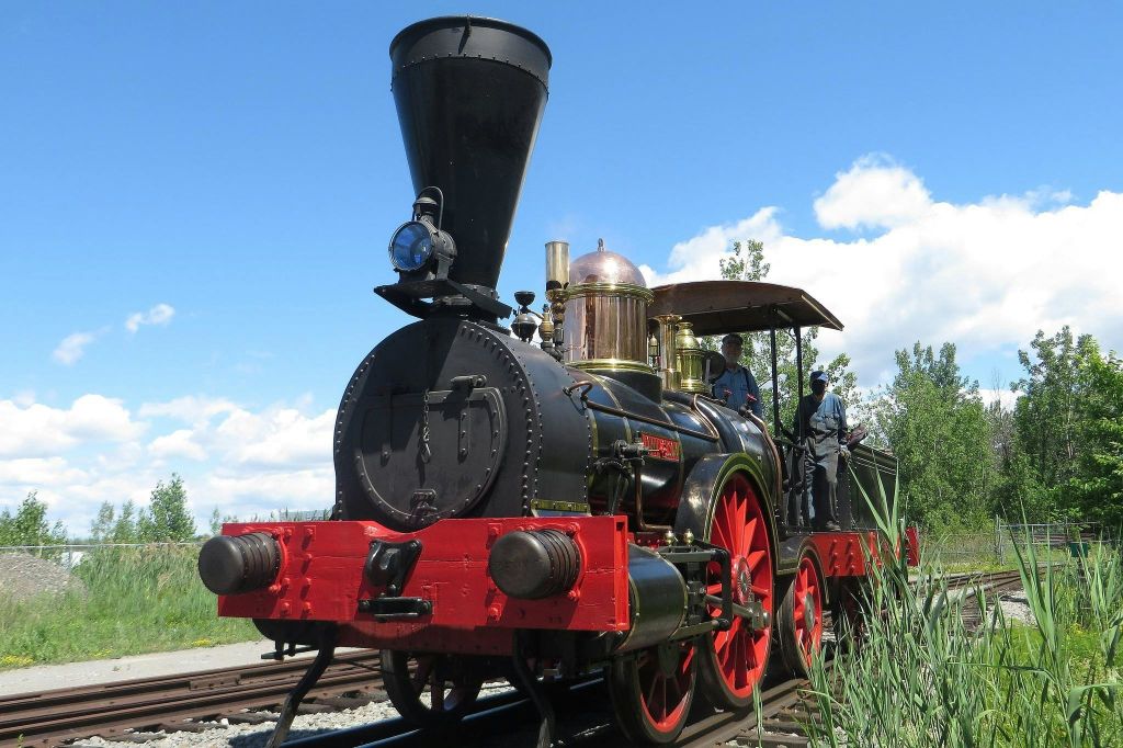 빨간색 바퀴와 황동 디테일이 있는 빈티지 증기 기관차가 철로를 따라 이동합니다. 택시에는 두 사람이 보입니다.