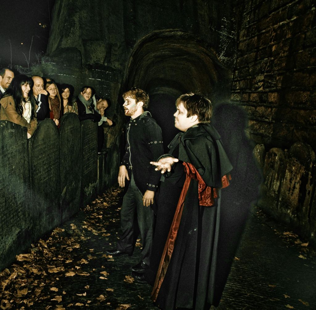 의상을 입은 공연자 두 명이 밤에 낙엽으로 덮인 낡고 돌담 옆에서 구경꾼들과 상호 작용합니다.