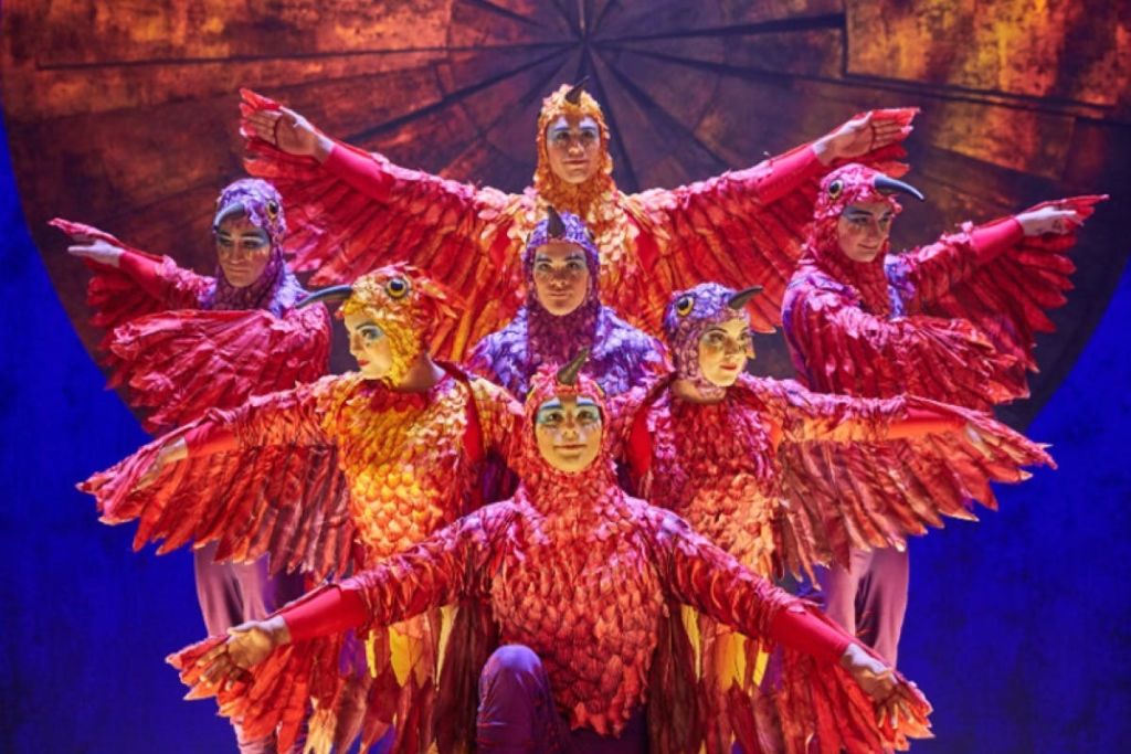 무대에서 따뜻한 조명 아래 펼쳐진 날개가 달린 화려한 새 의상을 입은 공연자들이 단체 포즈를 취하고 있습니다.