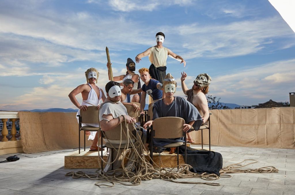 파란 하늘 아래 의자와 밧줄이 놓인 야외 세트에서 흰색 마스크와 다양한 의상을 입은 사람들이 포즈를 취하고 있습니다.